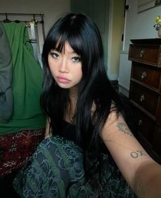 a woman with long black hair sitting on the floor next to a dresser and clothes rack