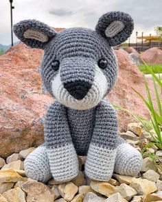 a crocheted gray and white teddy bear sitting on top of rocks next to grass