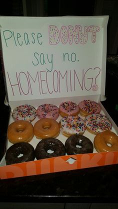 a box filled with lots of donuts sitting on top of a wooden table next to a sign that says donut say no home coming?