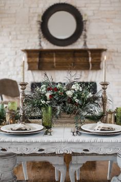 the table is set with candles, plates and flowers in front of a brick fireplace