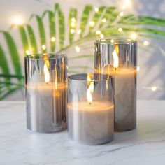 three lit candles sitting on top of a white table next to green plants and lights