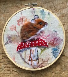 a small rodent sitting on top of a mushroom in a cross stitch hoop hanging from a piece of wood