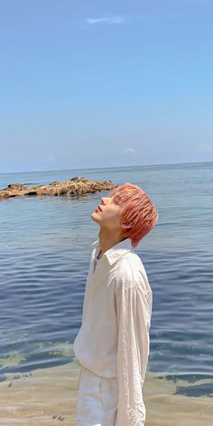 a man with pink hair standing on the beach looking up at the sky and water