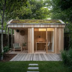 a wooden shed with a green roof and two chairs in the back yard, surrounded by trees