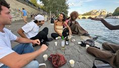 a group of people sitting on the side of a river next to each other eating and drinking