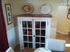 a white china cabinet with glass doors and plates on it's top, next to a dining room table