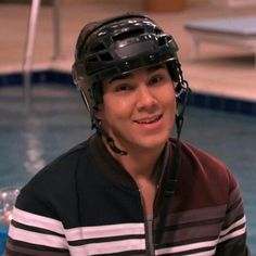 a young man wearing a hockey helmet sitting in front of a swimming pool and smiling at the camera