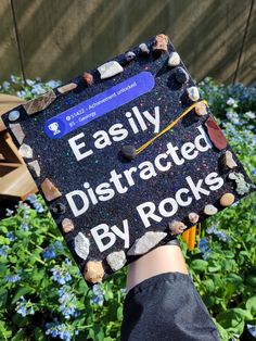 someone holding up a sign that says easily distracted by rocks in front of blue flowers