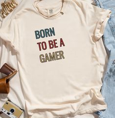 Shirt colors may vary slightly from what is viewed on computer screen. Unleash your inner gamer with our "Born to be a Gamer" t-shirt! Perfect for those who live and breathe video games, this shirt is a must-have for any true gaming enthusiast. Whether you're battling it out in the latest multiplayer game or reminiscing about classic retro titles, this t-shirt proudly declares your passion for all things gaming. Features: Premium Quality: Made from 100% soft, breathable cotton for ultimate comfo Gaming Event Crew Neck T-shirt With Letter Print, Gamer Style Cotton T-shirt With Letter Print, Gamer Style Short Sleeve Top With Letter Print, White Cotton Gamer T-shirt, Casual Letter Print Top For Gaming Events, Casual Tops With Letter Print For Gaming Events, Casual Top With Letter Print For Gaming Events, Casual Short Sleeve T-shirt For Gaming Events, Gamer Tops With Letter Print