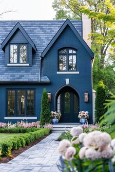 a blue house with lots of flowers in the front yard and bushes on either side