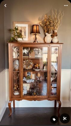 a wooden cabinet with glass doors and pictures on it's top shelf next to a lamp