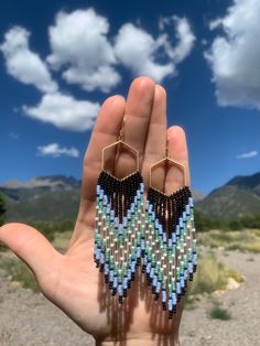 the hand is holding two pairs of beaded earrings in front of a blue sky with clouds