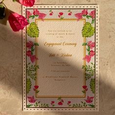 a wedding card with pink flowers and greenery on the front, sitting next to a red rose