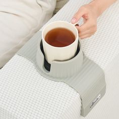 a person holding a cup of tea on top of a bed next to a pillow