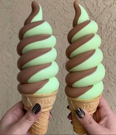 two ice cream cones with chocolate and mint green icing on their sides, held up in front of a stucco wall