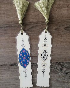 two tags with tassels hanging from them on a wooden surface, one is decorated with flowers and the other has an ornate design