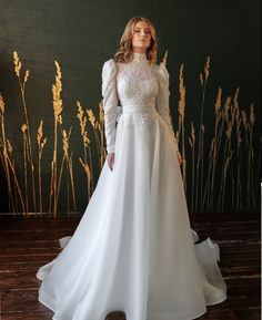 a woman standing in front of a wall wearing a white dress with long sleeves and lace