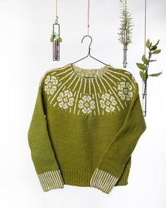 a green sweater with white flowers on it hanging from a hook and some plants in the background