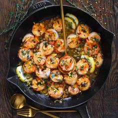 a skillet filled with shrimp and vegetables on top of a wooden table next to gold utensils