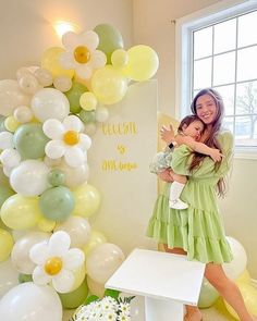 a woman holding a small child in her arms near balloons and flowers on the wall