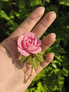 a hand holding a pink crocheted rose on it's palm with green leaves