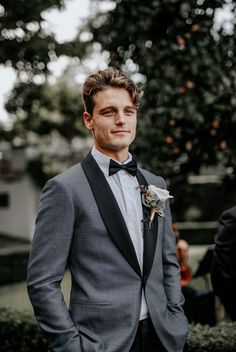 a man wearing a suit and bow tie standing in front of a hedged area