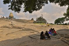 several people sitting on the rocks in front of a building and some trees with clouds overhead