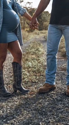 a pregnant woman holding the hand of a man in cowboy boots on a dirt road