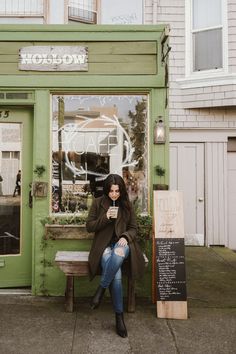 a woman sitting on a bench in front of a green building holding a coffee cup