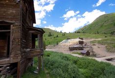 an old wooden house sitting on top of a lush green hillside next to a dirt road