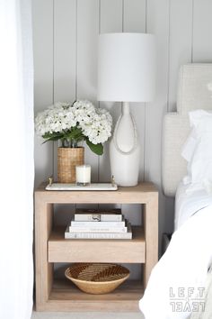 a nightstand with flowers and books on it next to a bed in a white bedroom