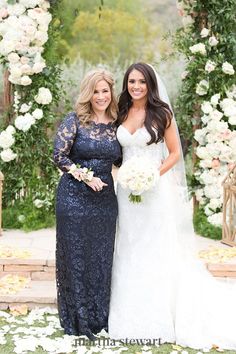 two women standing next to each other in front of a flower covered arbor with white flowers