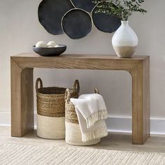 a wooden table topped with baskets next to a white vase and wall mounted art on the wall