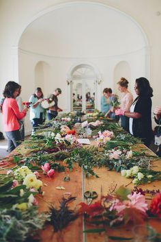 people standing around a long table with flowers on it and greenery in the middle