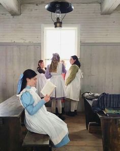 several women in white dresses are standing and sitting at desks, with one reading a book