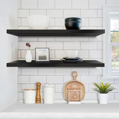 two black shelves with plates and bowls on them next to a window in a white kitchen