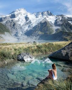 a woman sitting on top of a rock next to a body of water with mountains in the background