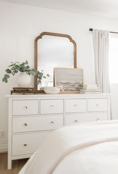 a white dresser with a mirror and vase on top of it next to a window