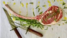 a raw piece of meat on a cutting board next to two knives and some herbs