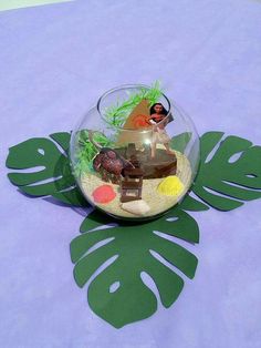 a fish bowl filled with plants and other items on a purple table cloth next to a green leaf