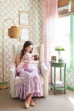 a woman holding a baby while sitting in a chair next to a window with pink and green wallpaper