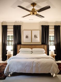 a large bed sitting under a ceiling fan in a bedroom next to two windows with black drapes