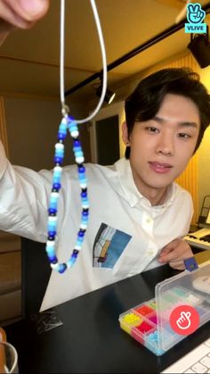 a young man wearing a white shirt and blue beaded necklace is standing in front of a computer