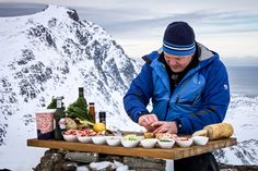 a man preparing food on top of a wooden table in the middle of snow covered mountains