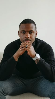 a man sitting on top of a couch wearing a black shirt and grey pants with his hands clasped