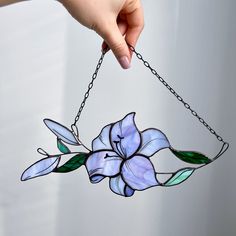 a hand holding a stained glass flower hanging from a chain with green leaves and purple flowers on it