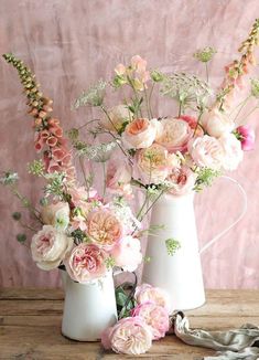 three white vases filled with pink flowers on top of a wooden table next to a cloth