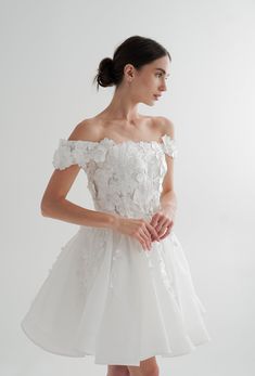 a woman wearing a white dress with flowers on the shoulders and shoulder, standing in front of