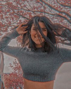 a woman standing in front of a tree with her hair blowing in the wind and wearing a gray sweater