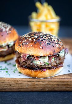 two burgers on a cutting board with fries in the background and text that reads blue cheese burgers on light broiled rolls / crispy panetta & onion chute